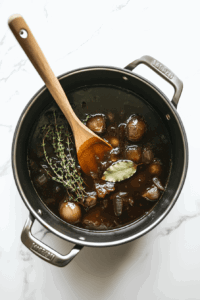 This image shows a soup pot being filled with caramelized onions, thyme, a bay leaf, and broth, combining ingredients for a hearty soup.
