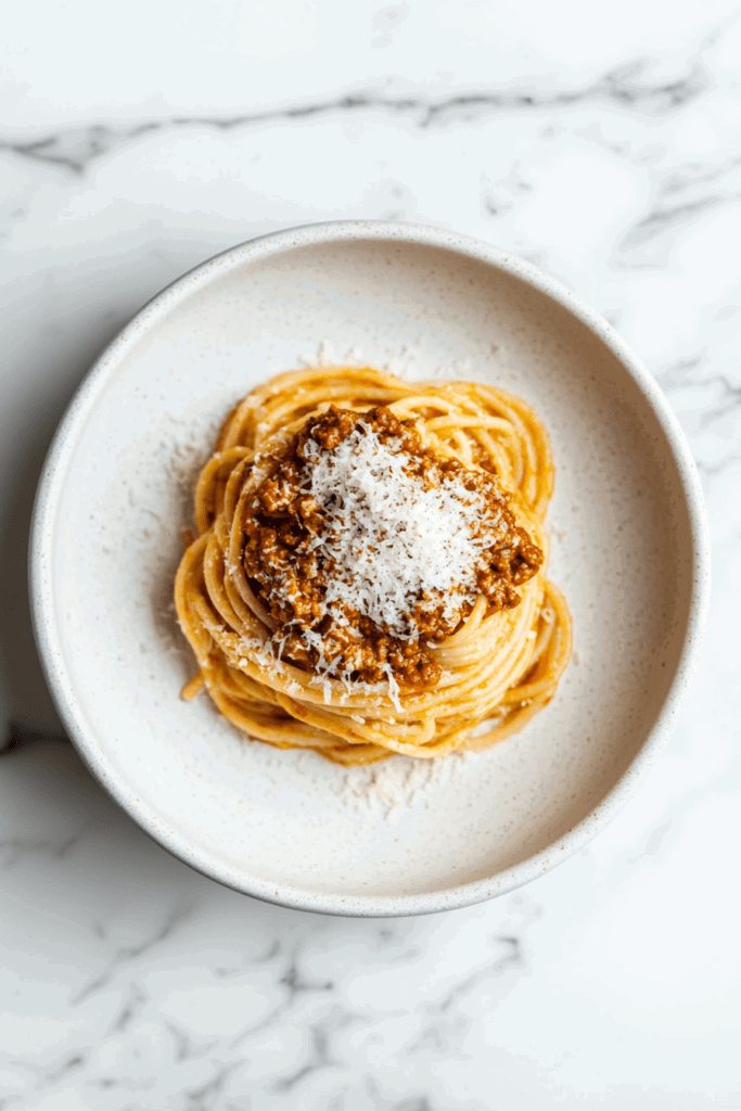 This image shows crockpot bolognese pasta served on a white round plate, topped with chopped meat and shredded cheese.