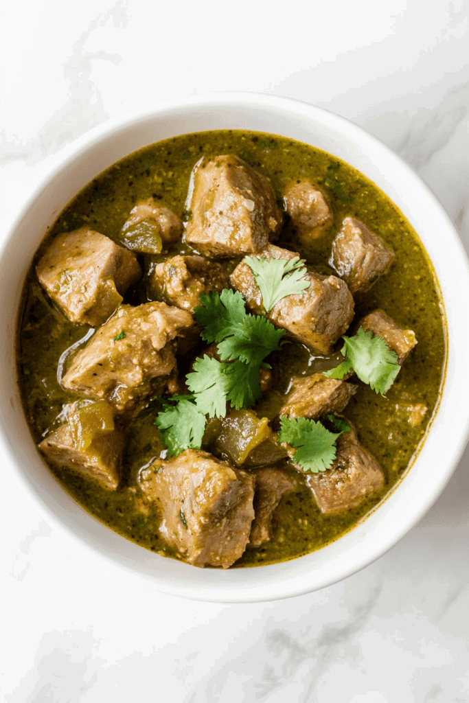 A zesty bowl of Crockpot Chili Verde with tender pork pieces and fresh cilantro leaves sprinkled on top, served in a simple white bowl.