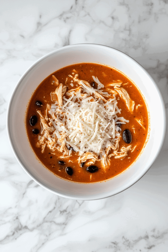 This image shows slow cooker chicken enchilada soup, a flavorful and hearty dish made with shredded chicken, beans, corn, and a rich enchilada sauce, garnished with fresh cilantro and served in a white bowl.