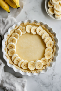 This image shows thinly sliced bananas being layered over the creamy filling in the baking dish, adding sweetness and texture to the pie.