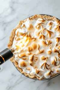 This image shows whipped aquafaba meringue being spread over the pie and lightly torched to create a golden, smoky topping for the vegan lemon meringue pie.