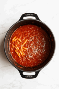 this image shows A pot filled with crushed tomatoes, vegetable broth, uncooked penne pasta, salt, and pepper, all combined to cook together in a one-pot pasta dish.