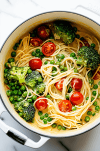 this image shows a vibrant mix of fresh broccoli, green peas, and cherry tomatoes being added to the pot, enhancing the one-pot vegan pasta primavera with color and flavor.