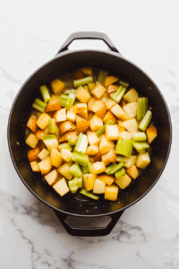 This image shows minced garlic, chopped celery, and diced potatoes being added to a Dutch oven with sautéed onions, building layers of flavor for the vegan corn chowder.