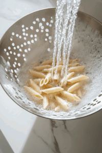 this image shows A large bowl of cooked penne pasta being mixed with vibrant green peas, sundried tomatoes, fresh arugula, cucumber slices, and creamy vegan pesto sauce.