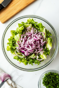 This image shows thinly sliced red onion rings being spread over the lettuce layer, adding a pop of vibrant purple color to the salad.