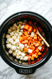 This image shows chopped sweet potatoes, cauliflower florets, and curry spices being added to a slow cooker, ready to be transformed into a creamy and aromatic soup.