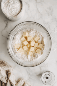 This image shows small cubes of vegan butter being added to the flour mixture in a food processor, preparing for blending.