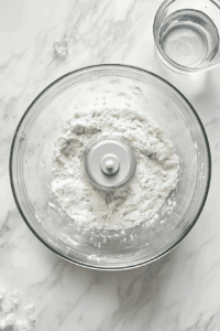 This image shows water being added to the processed ingredients in a food processor to form a cohesive dough for the pie crust.