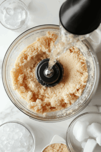 This image shows a small stream of ice-cold water being added to the flour and butter mixture in the food processor to create a cohesive dough.