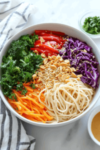 this image shows Cooked rice noodles arranged in a bowl and topped with colorful sliced vegetables, creating a visually appealing base for the Vegan Rainbow Peanut Noodles.