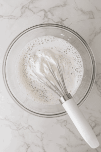 This image shows the chickpea liquid being whipped in a mixing bowl with a hand mixer, starting to form soft peaks for vegan whipped cream.