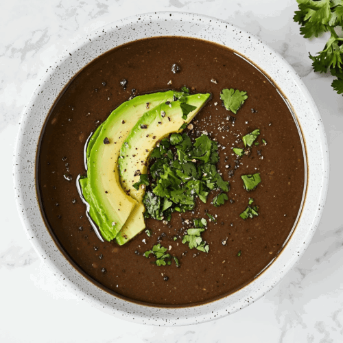 This image shows a hearty black bean soup served in a white bowl, topped with fresh tomato salsa, sliced avocado, and a sprinkle of fresh cilantro for a vibrant and flavorful finish.