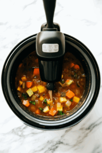 This image shows an immersion blender pureeing a mixture of sweet potatoes, cauliflower, and spices into a smooth and creamy curried soup directly in the slow cooker.