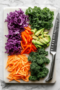 this image shows An array of vibrant vegetables, including carrots, red peppers, and cucumbers, being sliced and prepped for the Vegan Rainbow Peanut Noodles.