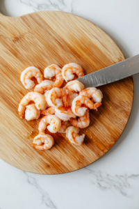 This image illustrates shrimp being chopped into small, bite-sized pieces on a cutting board, ready to be mixed into the shrimp salad topping.
