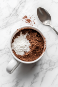 This image shows dry ingredients like flour, cocoa powder, sugar, and baking powder being added to a mug, preparing the base for a quick chocolate mug cake.