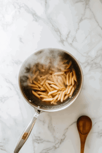 this image shows Penne pasta being cooked in a large pot of boiling water, preparing the base for the 6-ingredient vegan pesto pasta salad.