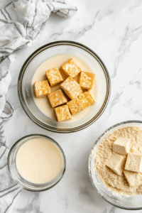 This image shows Marinated tofu pieces being dipped in vegan buttermilk and coated with a seasoned flour mixture, creating a crispy breaded layer.al.