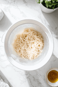 this image shows Cooked rice noodles being drained in a colander and rinsed under cold water to prevent sticking, preparing them for the Vegan Rainbow Peanut Noodles.