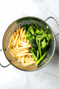 this image shows Cooked penne pasta, carrots, and broccoli florets being drained in a metal colander, ready to be combined with the sauce for Vegan Pasta Primavera.