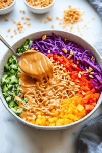 this image shows A drizzle of creamy peanut sauce being added to the Vegan Rainbow Peanut Noodles, garnished with crushed peanuts and sesame seeds for a flavorful and colorful presentation.