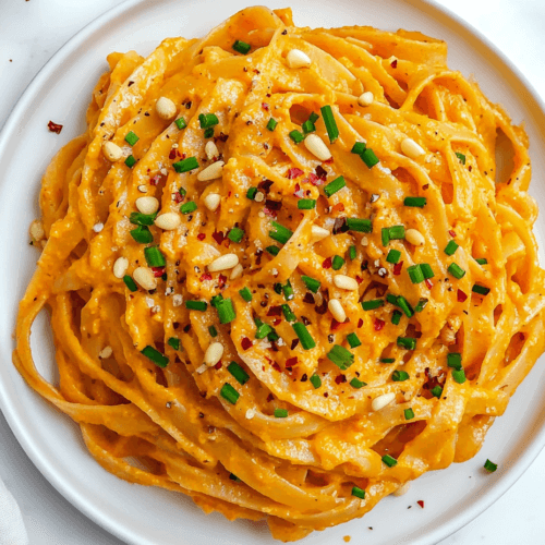 this image shows A beautifully plated serving of sweet potato fettuccine garnished with chopped spring onions, white sesame seeds, and chili flakes, served on a white round plate for a flavorful and vibrant meal.