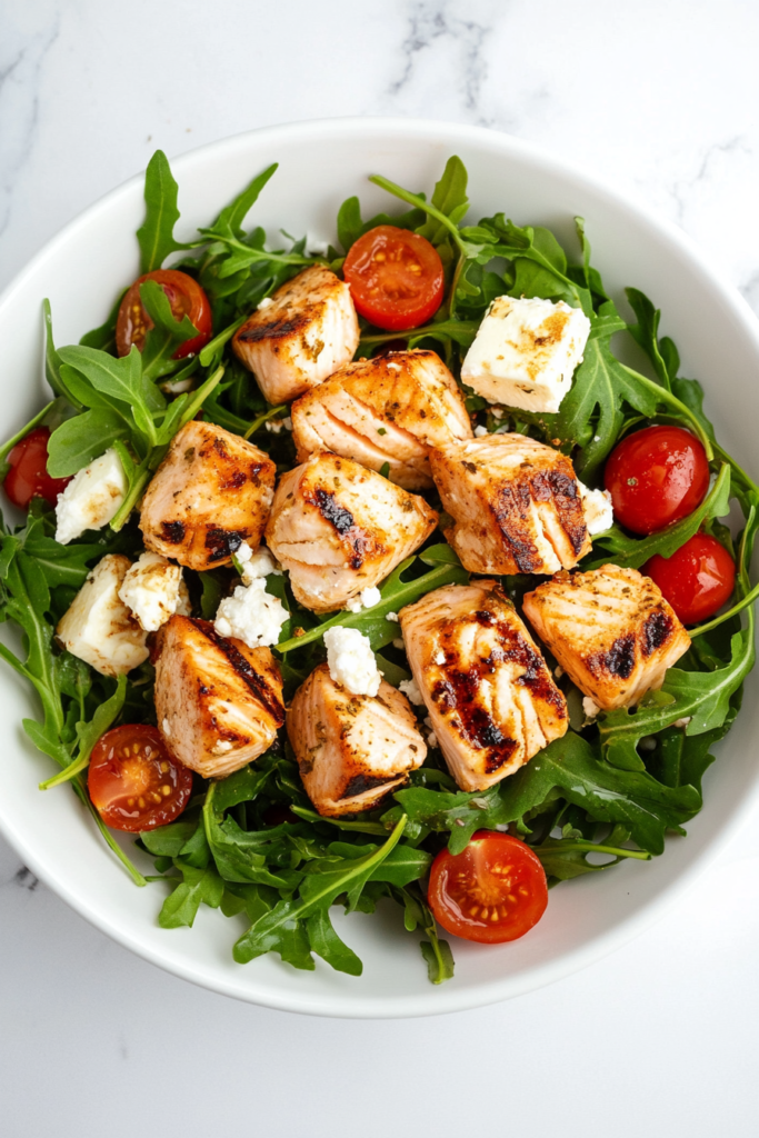 This image shows a white bowl of feta salmon salad, made with fresh spring mix greens, chopped cucumber, tomatoes, crumbled feta cheese, and perfectly cooked salmon pieces.