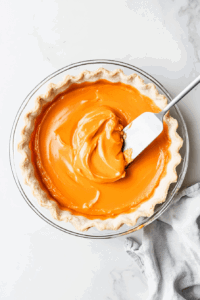 This image shows the silky pumpkin pie filling being poured into a pre-baked crust, spreading evenly to form the base of the vegan dessert.