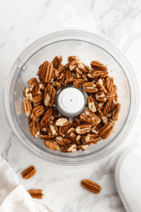 this image shows raw pecans being ground in a food processor to create a fine, crumbly texture, forming the base of the crust for the chocolate truffle pie.