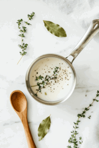 This image shows a saucepan with almond milk being gently heated alongside fresh herbs and aromatics, creating a flavorful base for vegan béchamel sauce.