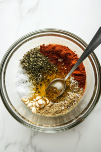 This image shows olive oil, garlic powder, oregano, basil, pepper, salt, onion powder, and Dijon mustard being combined in a large mixing container as the base for Greek salad dressing.