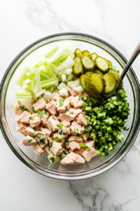 this image shows bowl of tuna, diced celery, chopped pickles, and finely chopped onions being gently stirred into the creamy dressing, creating a flavorful base for the tuna salad with egg.