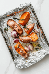 this image shows Sweet potatoes, shallots, and garlic cloves being chopped and placed on a baking sheet, ready to roast, while the oven preheats.