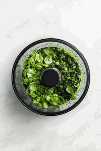 this image shows Fresh avocado, basil leaves, garlic, and seasonings placed in a food processor, ready to be blended into a creamy vegan pesto sauce for the pasta.