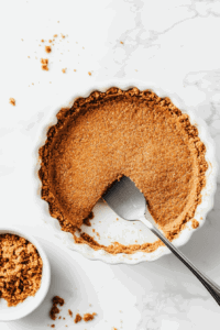 This image shows a ginger cookie crust mixture being firmly pressed into the bottom and sides of a pie dish to form the base for the vegan lemon pie, ready to be set in the freezer.