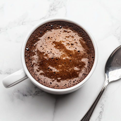 this image shows A delicious chocolate mug cake served in a white mug with a spoon placed beside it, showcasing a quick and satisfying dessert ready in just a few minutes.