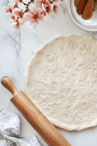This image shows dough being rolled out evenly with a rolling pin on a lightly floured surface, preparing it for fitting into the pie tin.
