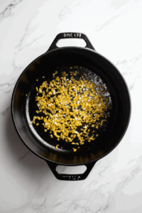 This image shows diced onions sizzling in a Dutch oven with a sprinkle of salt, creating a fragrant base for the creamy vegan corn chowder.