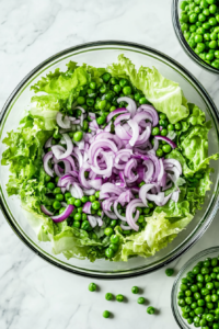 This image shows bright green peas being scattered evenly on top of the red onion layer, creating a colorful and sweet addition to the salad.