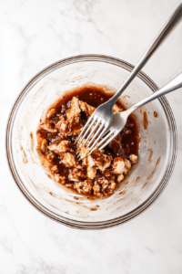 Fully cooked chicken breasts being shredded with a fork, creating tender pieces perfect for soaking up the BBQ sauce.