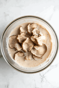 this image shows Oyster mushrooms soaking in a creamy vegan buttermilk mixture to absorb flavor and prepare for the flour coating.