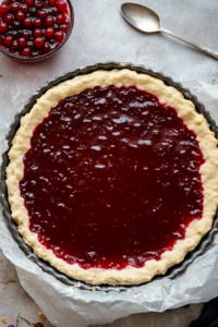 This image shows a rich layer of red currant jelly being evenly spread over the dough base, adding vibrant color and tart flavor to the Vegan Linzer Torte.