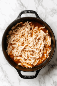 This image shows shredded chicken being stirred into a hot pan, evenly mixing it with other ingredients to enhance the flavor and texture of the dish.