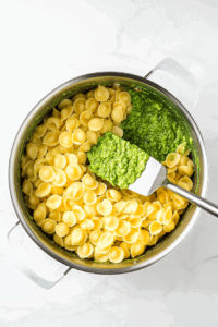 this image shows Hot, cooked pasta being tossed with creamy avocado pesto sauce in a large mixing bowl, ensuring each strand is coated with the vibrant green sauce.