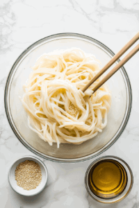 this image shows Rice noodles being tossed with sesame oil in a large bowl, adding a subtle nutty flavor and preventing clumping.