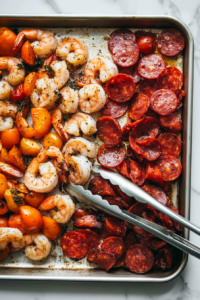 This image shows the ingredients on the sheet pan being tossed to evenly coat the shrimp and vegetables with the seasonings before roasting.