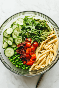 this image shows Tossing the vegan pesto pasta salad in a glass bowl, ensuring the pasta and vegetables are evenly coated with the flavorful pesto dressing.