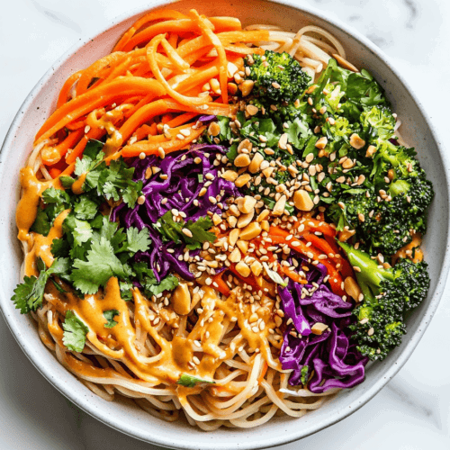 this image shows A white ceramic bowl filled with vibrant Vegan Rainbow Peanut Noodles, topped with colorful vegetables, crushed peanuts, sesame seeds, and a drizzle of creamy peanut sauce.